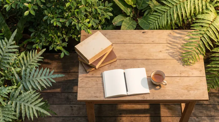 A stack of well-read books beside a cup of tea on a wooden table with green plants and morning light