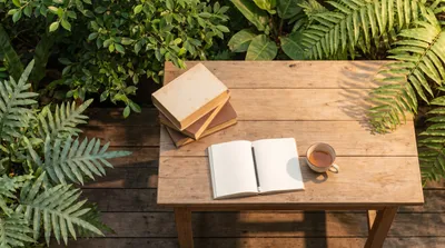 A stack of well-read books beside a cup of tea on a wooden table with green plants and morning light
